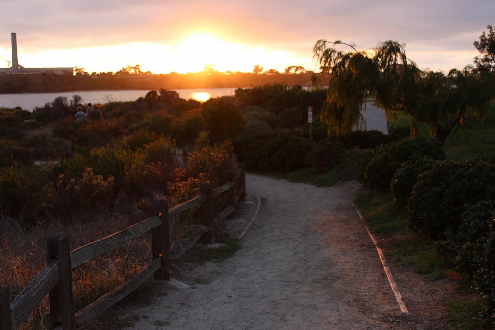Footpath beside Agua Hedionda Lagoon at sunset, with the low sun over the water and a wooden fence along the trail.