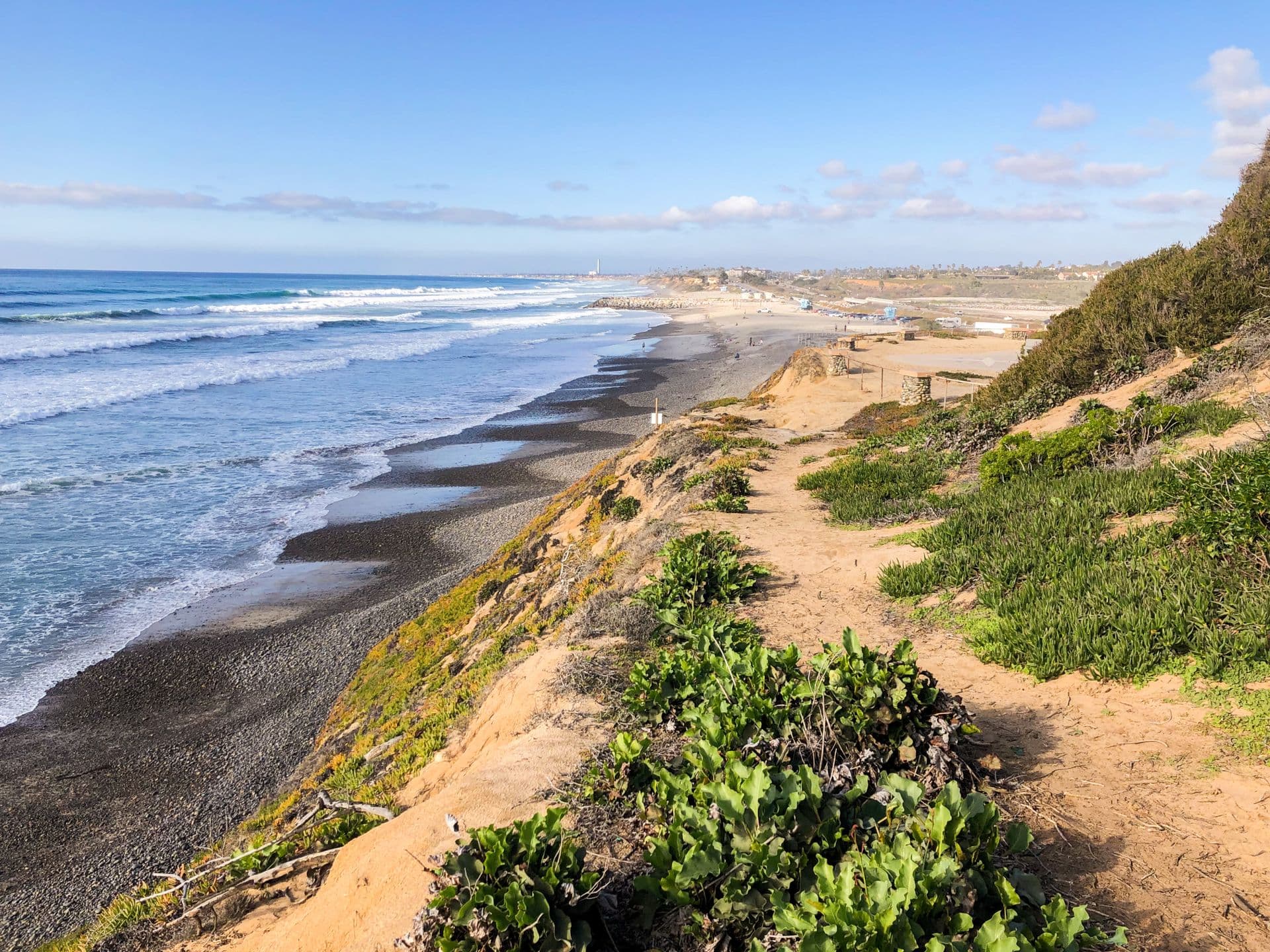 A sandy bluff trail above the Carlsbad coast, with surf rolling onto a long beach under a clear blue sky.