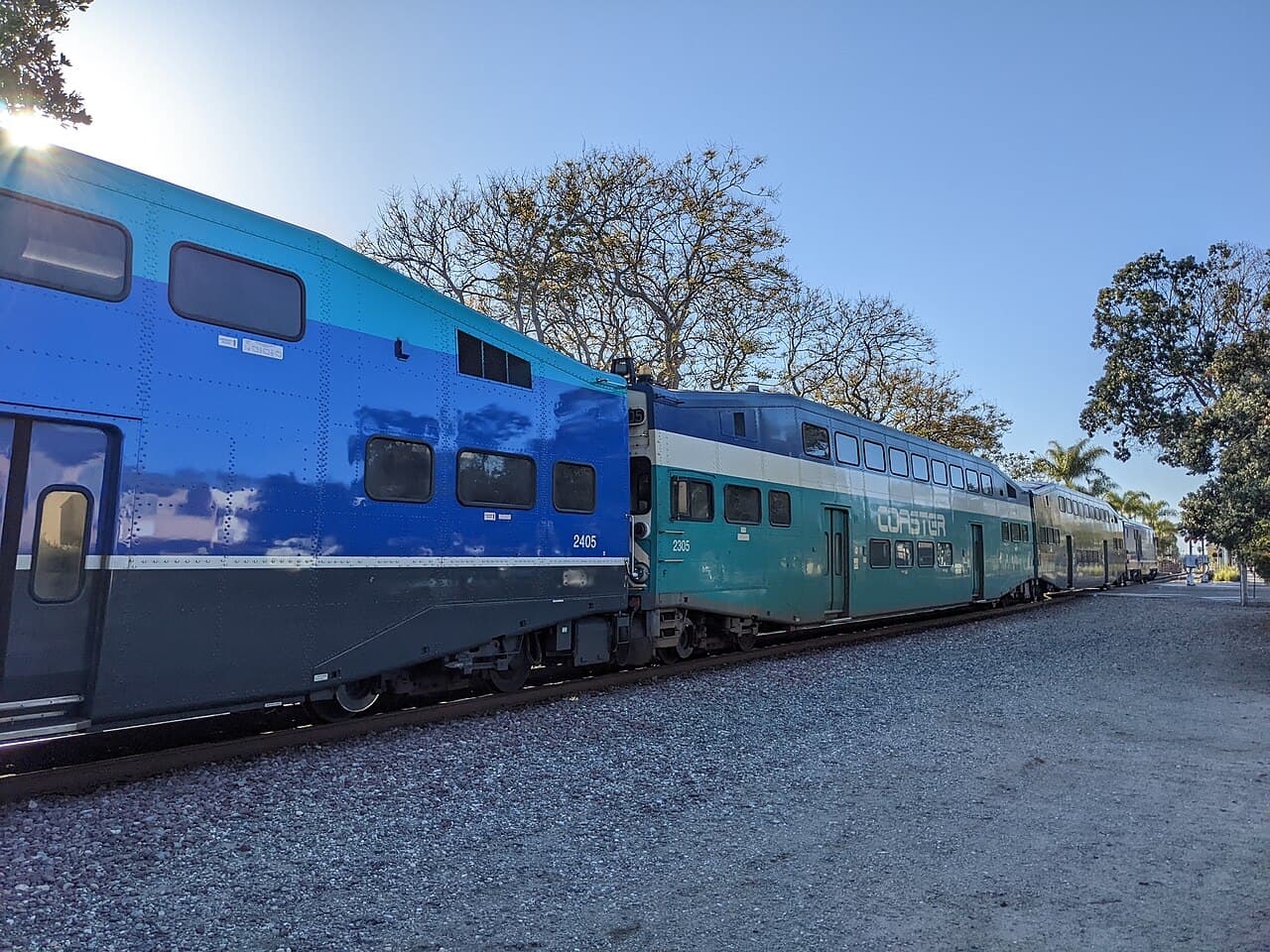 A blue and teal double-decker Coaster commuter train standing beside the gravel platform at Carlsbad Village, with trees behind.