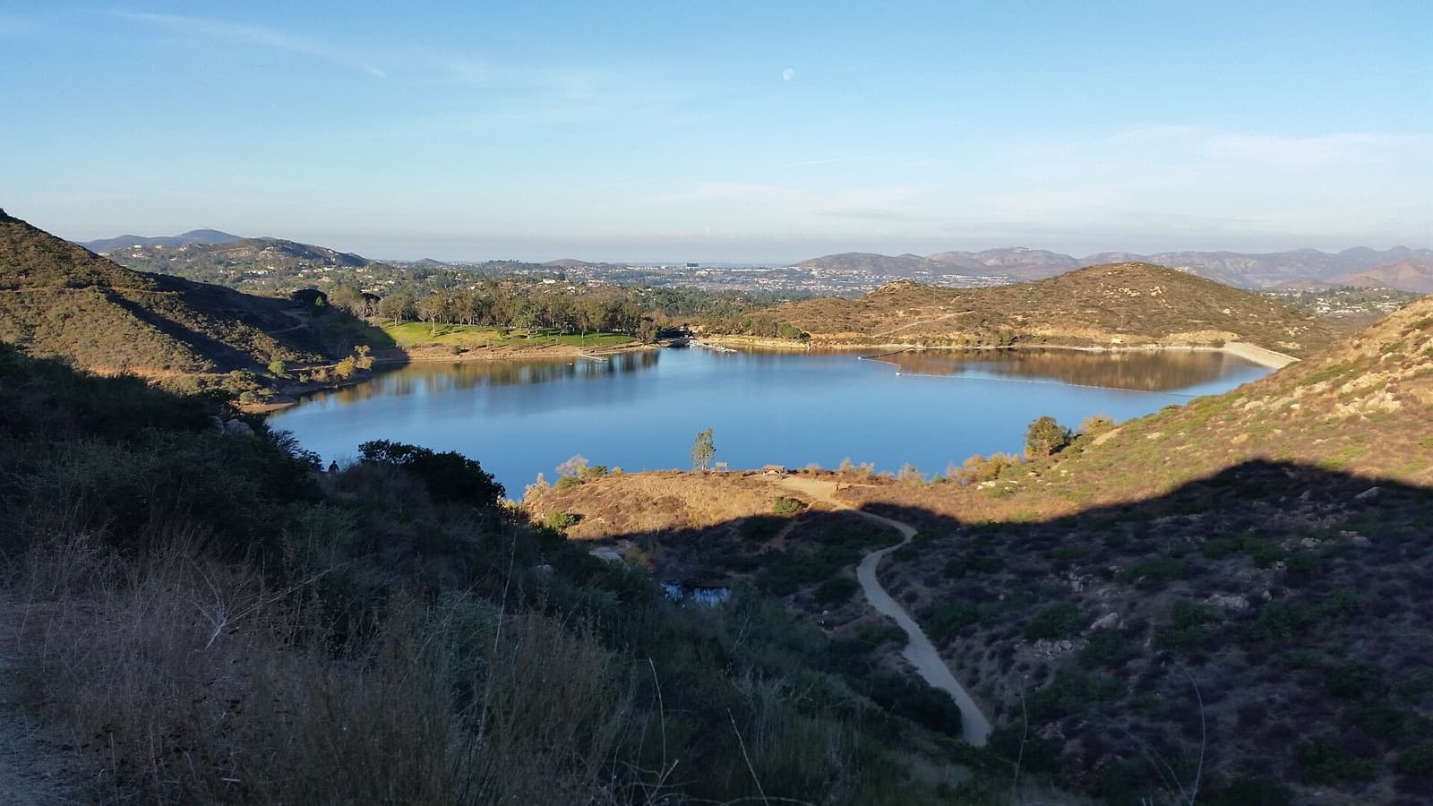 Lake Poway seen from a hillside trail in golden morning light, with a winding path below and hills beyond.