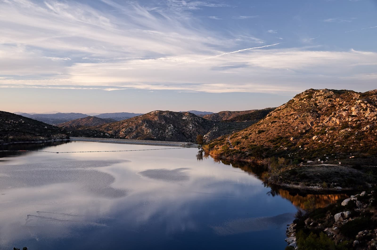 Calm water at Lake Poway reflecting a streaked evening sky, with the dam and rocky hills beyond.